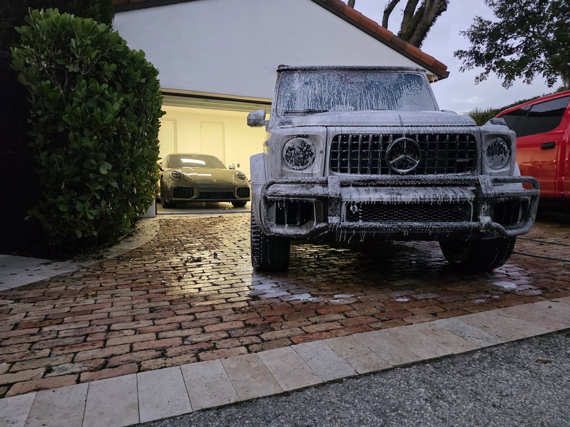 Mercedes G-Wagon covered in foam during a mobile detail at a home in Broward County, with a Porsche 911 in the lit garage behind it